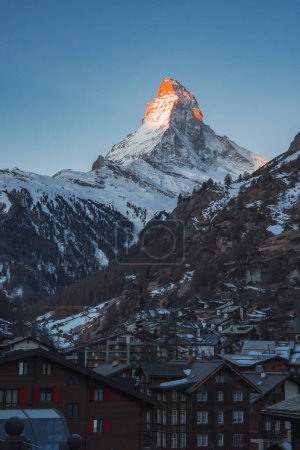 Heitere Dämmerung im Skigebiet Zermatt mit dem ikonischen Matterhorn in leuchtendem Orange. Schweizer Chalets umrahmen verschneite Landschaft und schaffen ruhige alpine Schönheit.