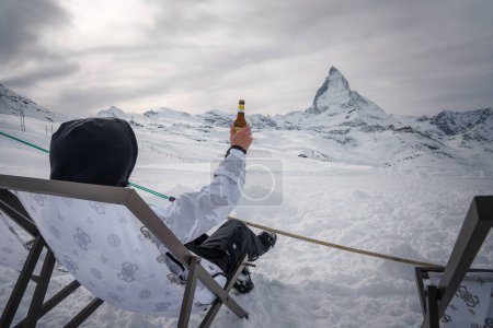 Escena de invierno en la estación de esquí de Zermatt persona en equipo de nieve sostiene la cerveza en la silla de cubierta con vistas a la montaña Matterhorn. Mezcla perfecta de aventura y relajación.