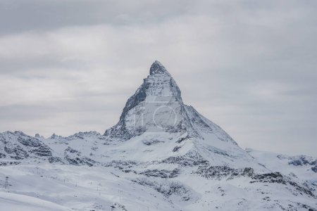 Matterhorn mountain, iconic Zermatt ski resort landmark in Switzerland. Distinctive pyramid peak against cloudy sky, snow covered slopes, ski lifts, trails. Natural alpine beauty.