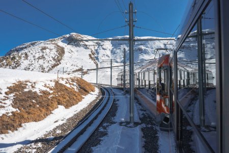 Train journey through Zermatt ski resort area, with scenic views from inside a white and orange carriage. Snow covered mountains and clear blue skies in the backdrop.