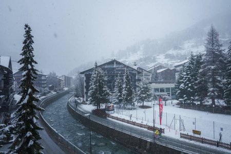 Photo for Snow covered Zermatt ski resort in Switzerland. River, red train, alpine buildings, evergreen trees, Swiss flag, and pedestrian in winter wonderland scene. - Royalty Free Image