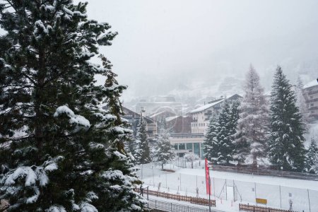 Verschneite Landschaft im Skigebiet Zermatt, Schweiz. Neuschnee auf immergrünen Bäumen, alpine Gebäude im traditionellen oder modernen Stil, im Vordergrund weht die Schweizer Flagge. Heitere Winterlandschaft.