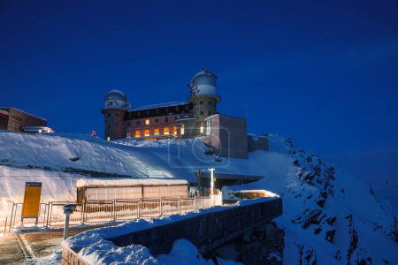Blue hour scene at high altitude Zermatt, Switzerland. Alpine building with observatory dome, snowy landscape, and starry sky. Ideal for winter travel and astronomy enthusiasts.