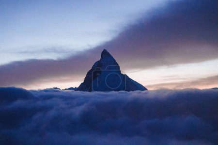 Iconic Matterhorn mountain in the Swiss Alps, silhouetted against a twilight sky with hues of blue and purple. Sea of clouds blankets the lower terrain. Serene atmosphere.