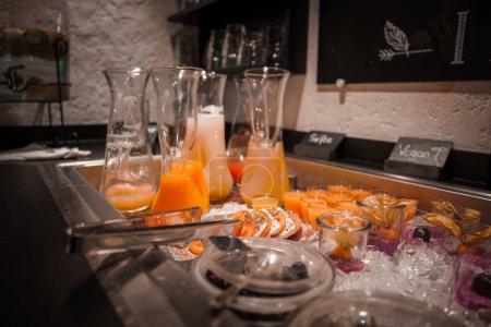 Büfett mit Glaskaraffen mit Orangensaft und kleinen Tassen im Luxushotel in Zermatt, Schweiz. Frische Obstschalen auf Eis, vegane Optionen verfügbar.