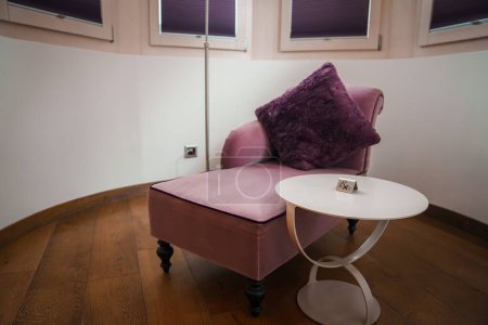 Luxus-Zimmerecke mit rosa Sessel, lila Kissen und weißem Tisch in Zermatt, Schweiz. Intimes, elegantes Dekor mit diffuser Beleuchtung zur Entspannung.