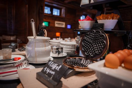 Breakfast buffet setup in a cozy wooden interior of a luxury hotel in Zermatt, Switzerland. Featuring a pot for hot beverages, waffle iron, condiments, and eggs. Ideal for post ski breakfast.