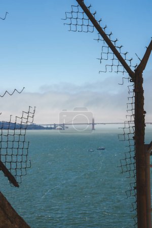Blick auf einen verrosteten Metallzaun am Alcatraz Gefängnis in Alcatraz Island, San Francisco Bay, CA, USA. Dahinter liegt die Bucht mit Booten und der Golden Gate Bridge im Hintergrund.