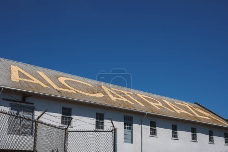 Nahaufnahme eines weißen Gebäudes im Alcatraz Gefängniskomplex in San Francisco, USA. ALCATRAZ in fetten gelben Buchstaben auf dem Dach, Maschendrahtzaun mit Stacheldraht im Vordergrund. Der Himmel ist klar blau.