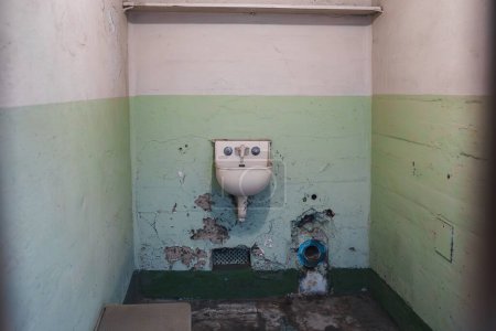 Abandoned room corner in Alcatraz prison. Peeling paint in white and faded green, exposed plumbing, damaged walls, worn floor. Evokes historical neglect.