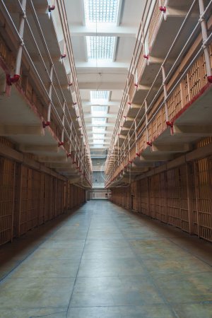 Interior view of Alcatraz prison, San Francisco, USA. Ground level perspective of cell block with barred doors, red pipes, worn green concrete floor, and windows for natural light.