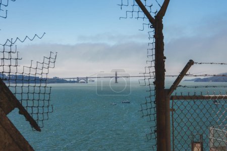 View through rusted bars and wire mesh inside Alcatraz overlooking San Francisco Bay. Golden Gate Bridge visible in distance with boats on calm water.