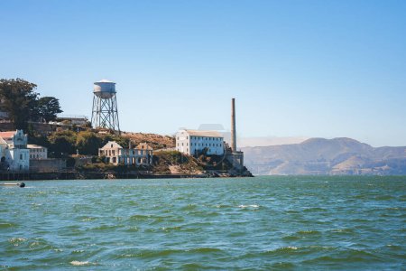 Scenic view of Alcatraz Island in San Francisco Bay, California, USA. Features former prison building, water tower, and coastal hills under blue sky. Peaceful waters.