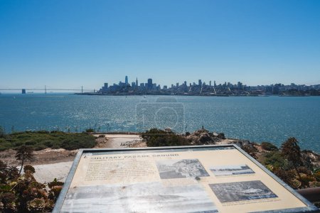 Scenic view of Alcatraz Island with MILITARY PARADE GROUND plaque, looking towards San Francisco skyline. Bay Bridge, boats visible. Sunny day, clear sky, no people.