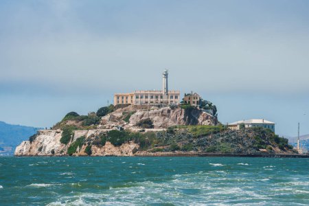 Alcatraz Island in San Francisco Bay, California, USA, viewed from the water. Shows rugged terrain, prison building, lighthouse, and choppy water under clear sky.