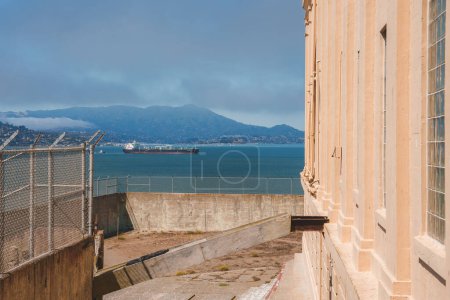 View from Alcatraz prison in San Francisco, USA. Image shows weathered beige exterior wall, barred windows, chain link fence, water, hilly landmass, cargo ship.