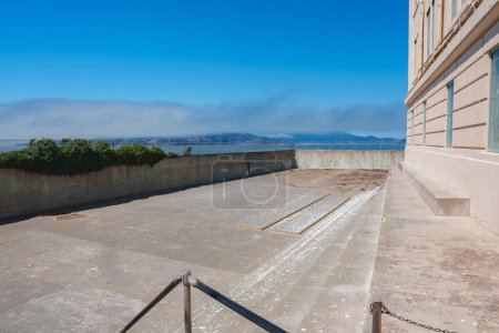 Outdoor area at Alcatraz prison in San Francisco, USA. Concrete pathway, metal railing, aged ground, barred windows, enclosed wall, lush foliage, scenic sky.