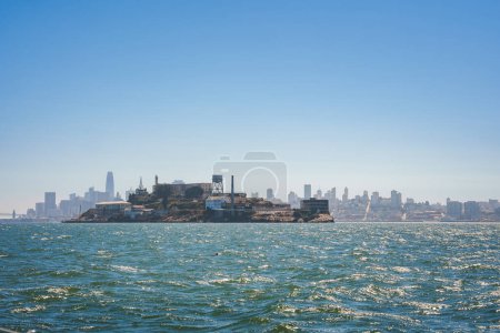 Scenic view of Alcatraz Island in San Francisco Bay under a clear sky, surrounded by old prison structures, guard towers, with city skyline in the background.