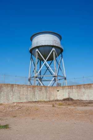 Tall, white water tower in Alcatraz, San Francisco. Metal beams form structure, encased in concrete wall with barbed wire. Neglected, arid environment.