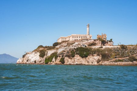 Alcatraz Island, San Francisco Bay, California, USA. Former federal prison perched on rocky island, with lighthouse. Rugged terrain, clear sky, calm waters.