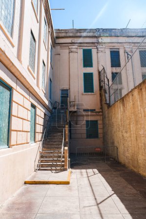 Alcatraz prison outdoor area with a narrow walkway, high walls, barred windows, metal staircase, teal shutters, chair, and bright daylight in San Francisco, USA.