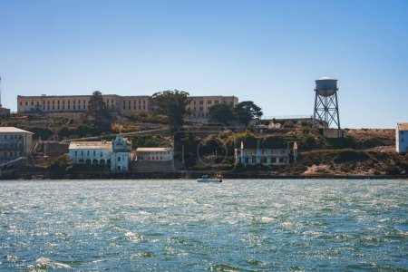 Clear day view of Alcatraz Island in San Francisco Bay. Image shows main prison building, other structures, rugged terrain, blue waters, and water tower.