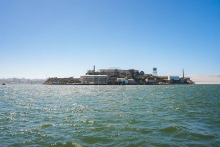 Scenic view of Alcatraz Island in San Francisco Bay on a sunny day. Shows the former prison complex with main cellhouse, water tower, and skyline in the distance.