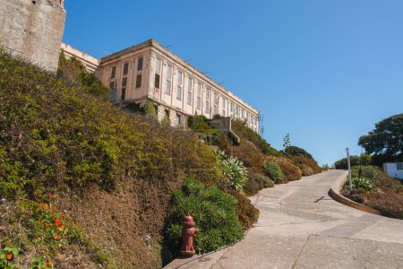 Exterior view of Alcatraz prison in San Francisco, USA. Large beige building atop hill with barred windows. Path, greenery, fire hydrant in foreground. Serene atmosphere with somber history.