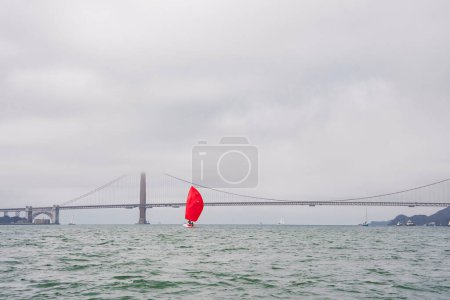 Serene view of San Francisco Bay with the Golden Gate Bridge covered by mist. A vibrant red sail yacht contrasts the calming water and muted sky colors. Ideal maritime scene.