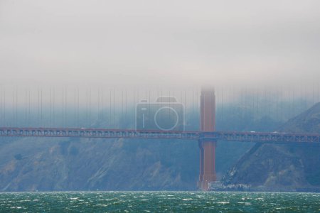 Iconic Golden Gate Bridge in San Francisco seen through foggy haze. Vibrant orange against blues and greens, choppy waters in foreground. Mystical view across the bay.