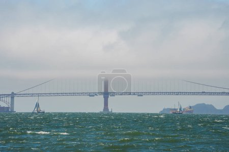 Iconic Golden Gate Bridge in mist, San Francisco. View from water shows bridge amidst choppy bay waters with sailboats. Overcast sky, hills in background. Shows natural beauty and engineering marvel.