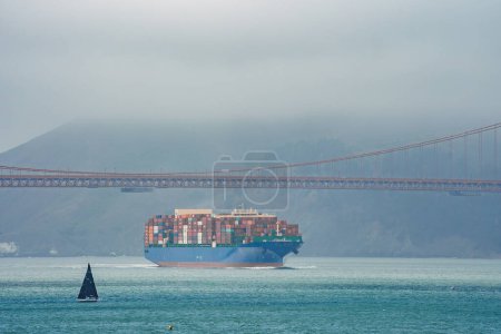 Ikonische Golden Gate Bridge in San Francisco Segelboot mit schwarzem Segel kontrastiert Frachtschiff mit bunten Containern gefüllt. Kühles, nebliges Wetter bringt Mystik in die Szene.