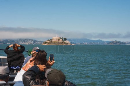 Foto de Los turistas en barco fotografian la isla de Alcatraz con teléfonos, contra el paisaje urbano de San Francisco. Traje casual, cielo nublado, serena vista al mar. Probablemente tomado de un barco de turismo. - Imagen libre de derechos
