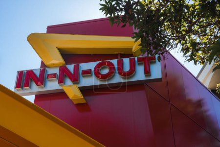 Icône rouge et blanc In N Out Burger signe sur fond rouge vif, détail jaune. Branche d'arbre, ciel bleu suggère une journée ensoleillée. Emplacement possible à San Francisco.