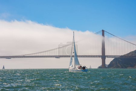 Iconic Golden Gate Bridge in San Francisco shrouded in fog with sailboats gliding on the Bay, reflecting sunlight. Hills and modern engineering in background.