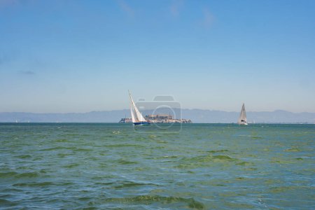Tranquil view of San Francisco Bay with choppy waters, sailboats, Alcatraz Island silhouette, and coastal mountains in the background, capturing the bays beauty and historical significance.