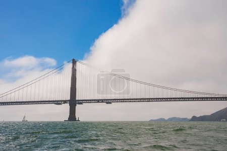 Szenische Ansicht der berühmten Golden Gate Bridge in San Francisco, CA. Turm, Hängeseile, Fahrzeuge auf dem Brückendeck, Segelboot auf dem Wasser, hügelige Küste in der Ferne.