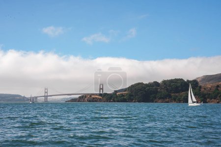 Serene view of Golden Gate Bridge over San Francisco Bay. White sailboat glides on calm water, hills covered in greenery, clear sky with scattered clouds.