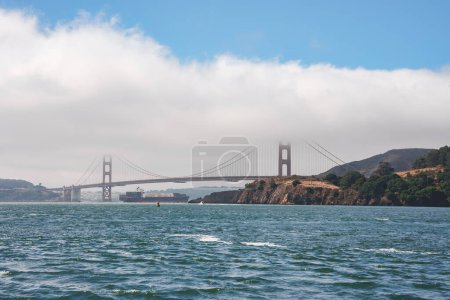 Tagesansicht Golden Gate Bridge, San Francisco, CA. Die symbolträchtige orangefarbene Brücke thront über abgehackten Gewässern, darunter fährt ein Frachtschiff. Majestätische, heitere Schönheit.