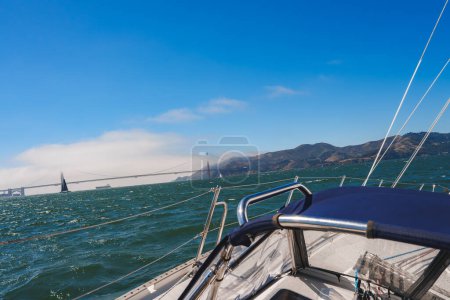 Yacht sailing near San Francisco, view from stern to bow with bridge, fog, sailboats, and green waters. Marin Headlands in background, clear sky overhead.