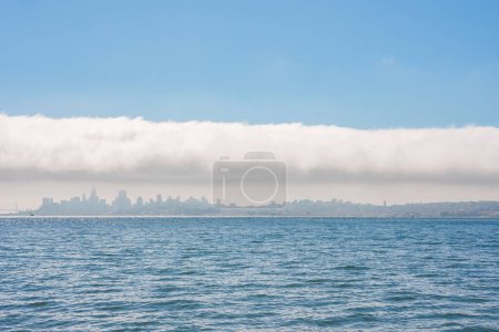 Discover the serene San Francisco skyline shrouded in low lying clouds, reflected in calm waters, under a clear sky. Iconic landmarks await in this picturesque cityscape.
