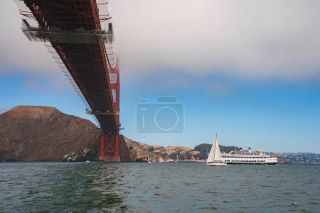 Iconic Golden Gate Bridge from below, towering red structure against blue sky in San Francisco Bay. Yacht sailing leisurely, Marin Headlands backdrop.