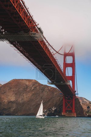 Ikonische Golden Gate Bridge in San Francisco, Kalifornien. Dramatischer Blick von unten mit hoch aufragender roter Stahlkonstruktion, nebligem Hintergrund. Segelboot gleitet anmutig durch ruhige Bucht.