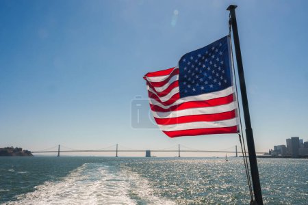 Téléchargez les photos : Drapeau américain flottant contre le ciel bleu, Golden Gate Bridge visible en arrière-plan sur la baie de San Francisco. Skyline de la ville faiblement visible à gauche. Composition fière. - en image libre de droit