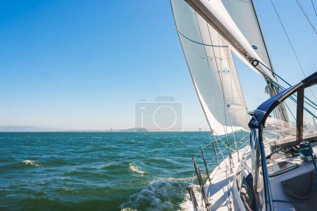 Sailboat sailing on serene waters near San Francisco, white sails billowing under a clear blue sky. Modern yacht deck and rigging visible, Bay Bridge in background.