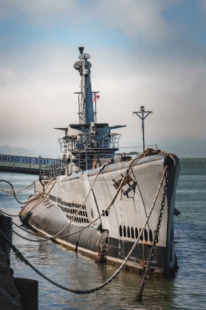 Photo for Large naval submarine with a dark grey hull and conning tower, moored at a pier, likely in San Francisco. Chains secure the vessel, under a partly cloudy sky. - Royalty Free Image