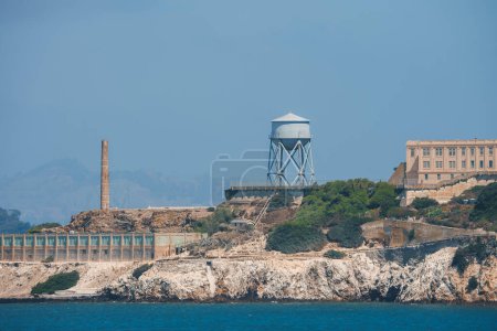 Distant view of Alcatraz Island in San Francisco Bay with rugged terrain, historical structures, water tower, and cellhouse. Popular tourist destination.