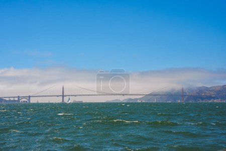 Vue panoramique du Golden Gate Bridge, San Francisco. Vagues dans la baie, brouillard, voiliers, ciel clair avec nuages, collines ensoleillées encadrent le monument emblématique.