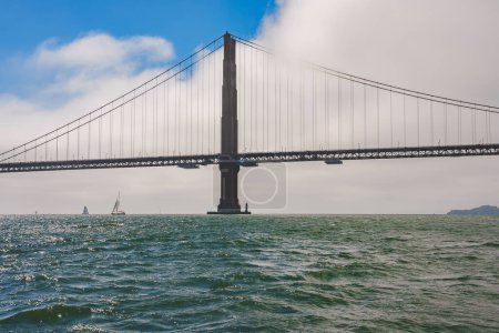Magnificent Golden Gate Bridge view in San Francisco, California, from water level. Iconic bridge tower, cables, sailboats, and foggy backdrop. Land on horizon.