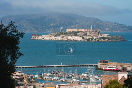 Malerischer Blick auf San Francisco mit der Insel Alcatraz im Hintergrund, geschäftiger Yachthafen mit Yachten, Segelboot auf dem Wasser, roter Sightseeing-Bus, Hügel in der Ferne.